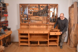 Mann öffnet Schublade an großer Werkbank mit geöffneter Werkzeugwand voller Handwerkzeuge.