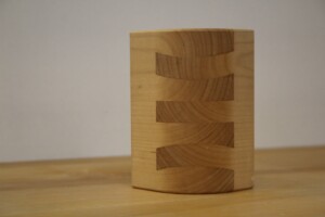 Holzstück mit geschwungener Fingerzinkung, stehend auf einer Holzoberfläche vor grauem Hintergrund.