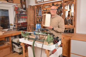 Mann in Werkstatt hält Holzkasten mit Schwalbenschwanzverbindung vor Fräsmaschine, lächelnd in Kamera.